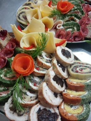 Dekoracyjnie ułożona zimna płyta z roladami mięsnymi, serami i wędlinami, ozdobiona pomarańczowymi kwiatami z warzyw i gałązkami kopru.