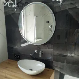 Nowoczesna łazienka z owalną umywalką nablatową, podświetlanym okrągłym lustrem i czarno-białymi płytkami imitującymi marmur, oświetlona dekoracyjnymi kinkietami.