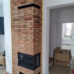Leszek Terka - Murowany kominek z cegły w trakcie budowy, zabezpieczony taśmą malarską, z widocznym wkładem kominkowym i otworami wentylacyjnymi, na podłodze folia ochronna.