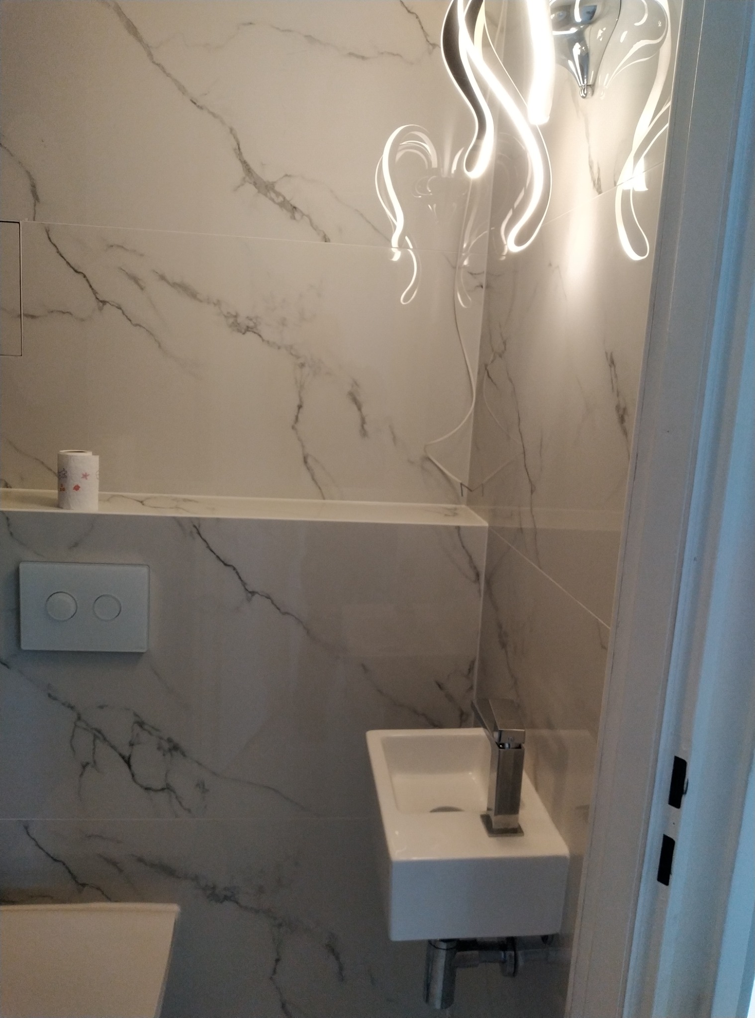 Nowoczesna łazienka z marmurowymi płytkami, mała umywalka z chromowaną baterią, designerska lampa sufitowa z białego szkła, widoczny fragment toalety i włącznik światła.