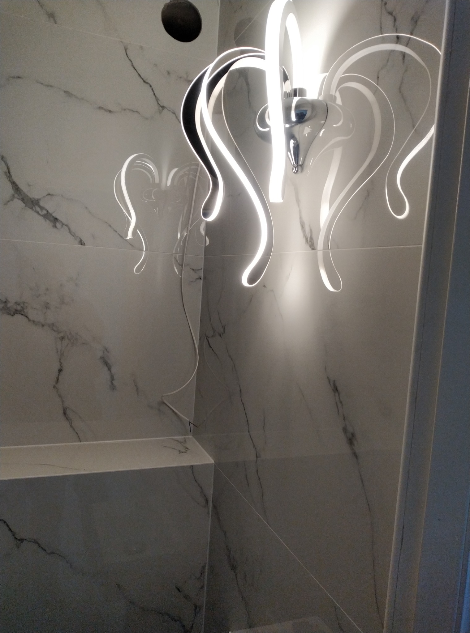 Nowoczesna lampa LED z elementami chromowanymi, zamontowana w łazience z marmurowymi płytkami, widoczny otwór w suficie na instalację.