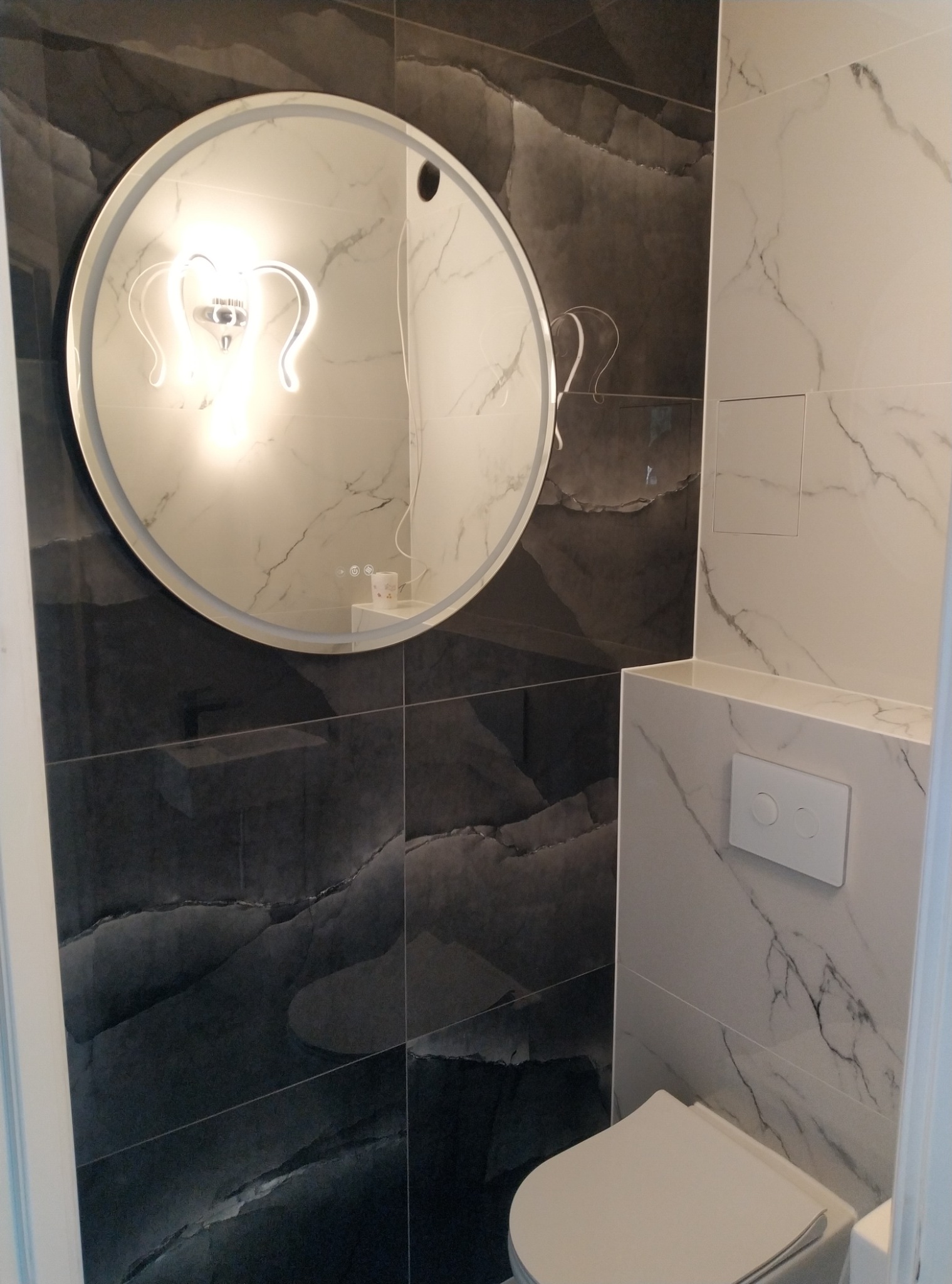 Wykończona łazienka z czarno-białymi płytkami imitującymi marmur, okrągłe lustro z podświetleniem LED w kształcie serca oraz podwieszana toaleta z białym przyciskiem spłukującym.