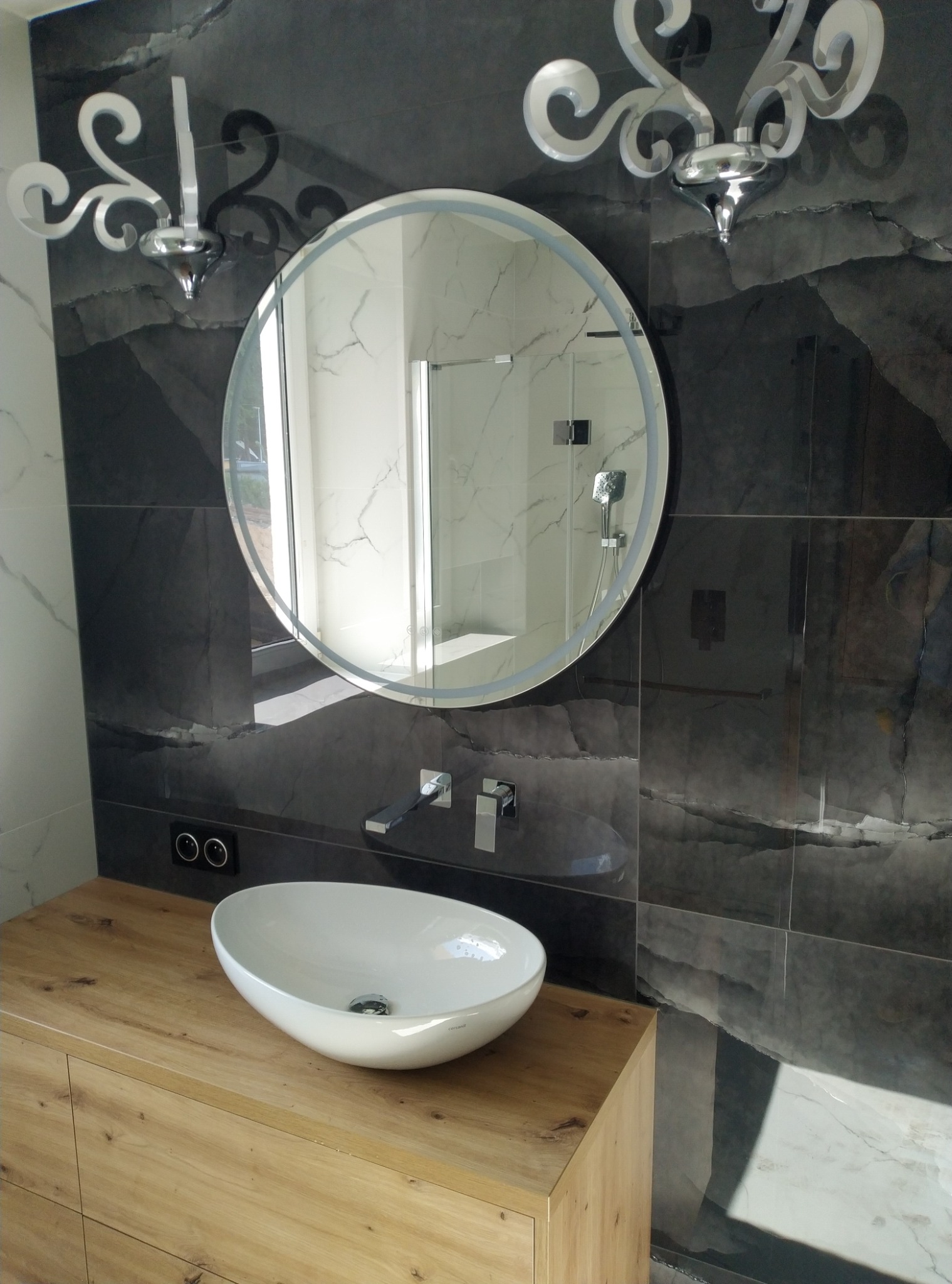 Nowoczesna łazienka z owalną umywalką nablatową, podświetlanym okrągłym lustrem i czarno-białymi płytkami imitującymi marmur, oświetlona dekoracyjnymi kinkietami.