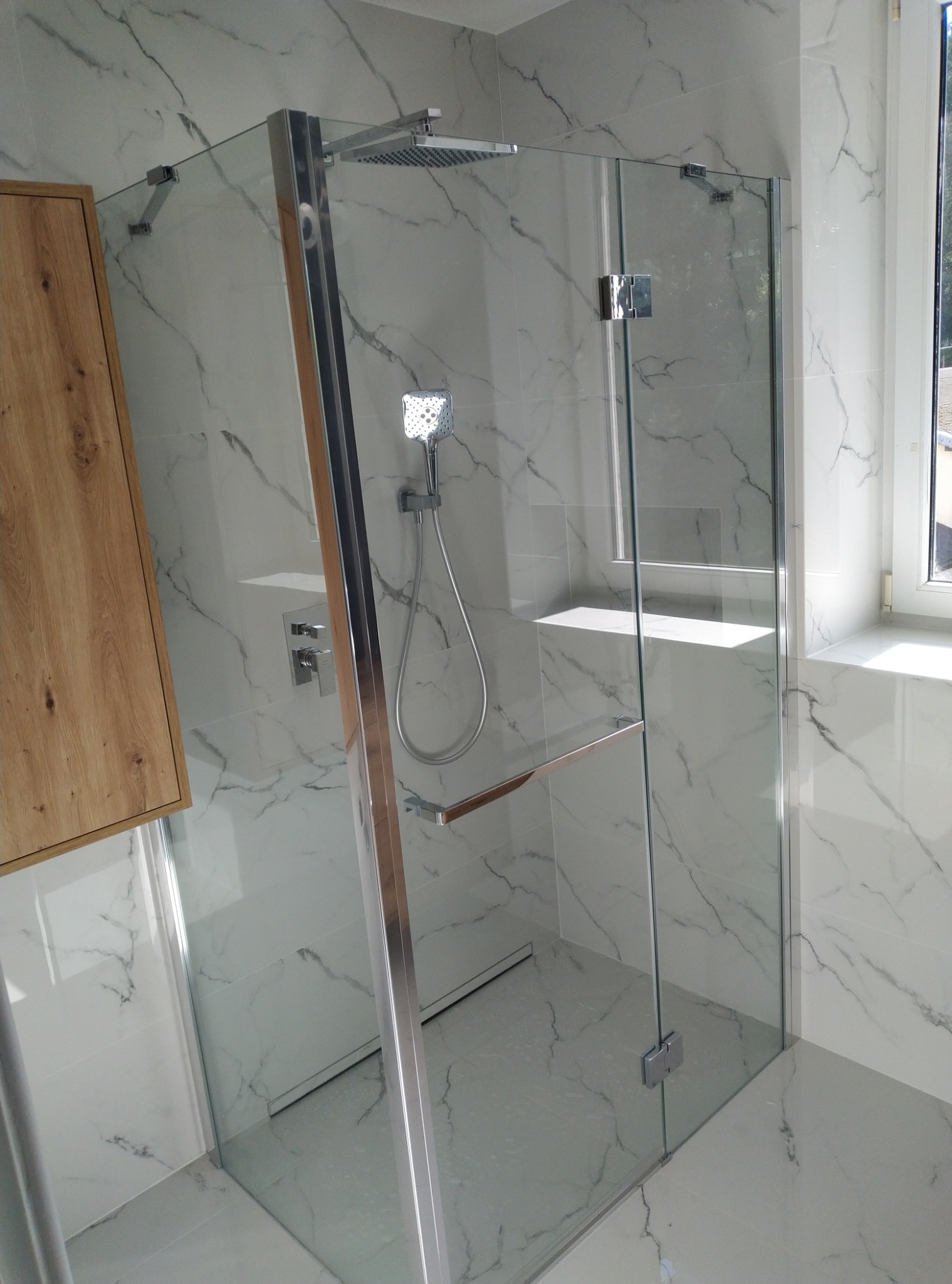 Nowoczesna kabina prysznicowa z chromowanymi okuciami i deszczownicą, wykończona białymi płytkami imitującymi marmur, z drewnianą szafką w tle.