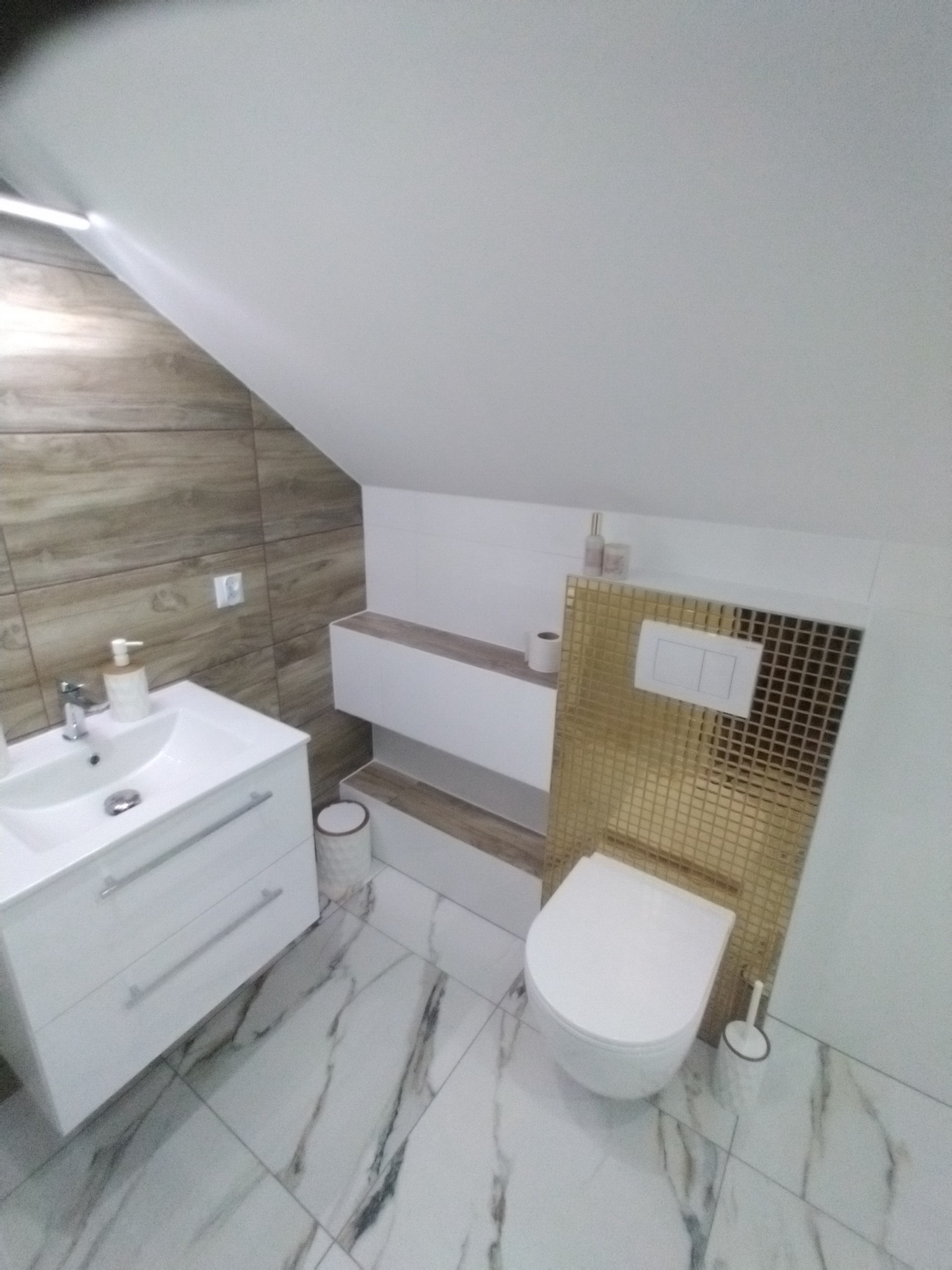 Nowoczesna łazienka na poddaszu z umywalką na szafce, toaletą z mozaiką i marmurową podłogą.