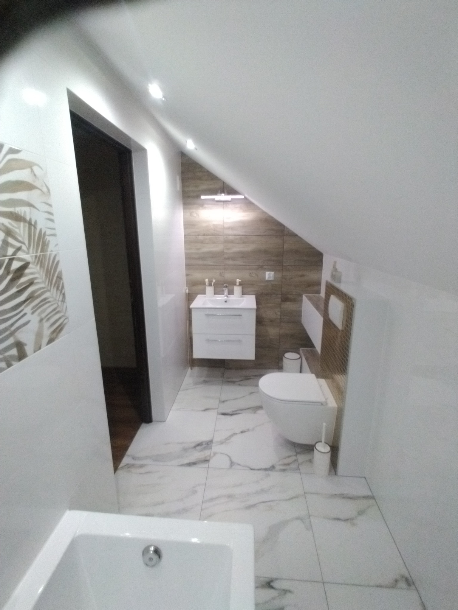 Wykończona łazienka na poddaszu ze skośnym sufitem, białe płytki z marmurowym wzorem na podłodze, umywalka z szafką w kolorze białym, toaleta typu geberit, ściana z brązowych płytek imitujących...