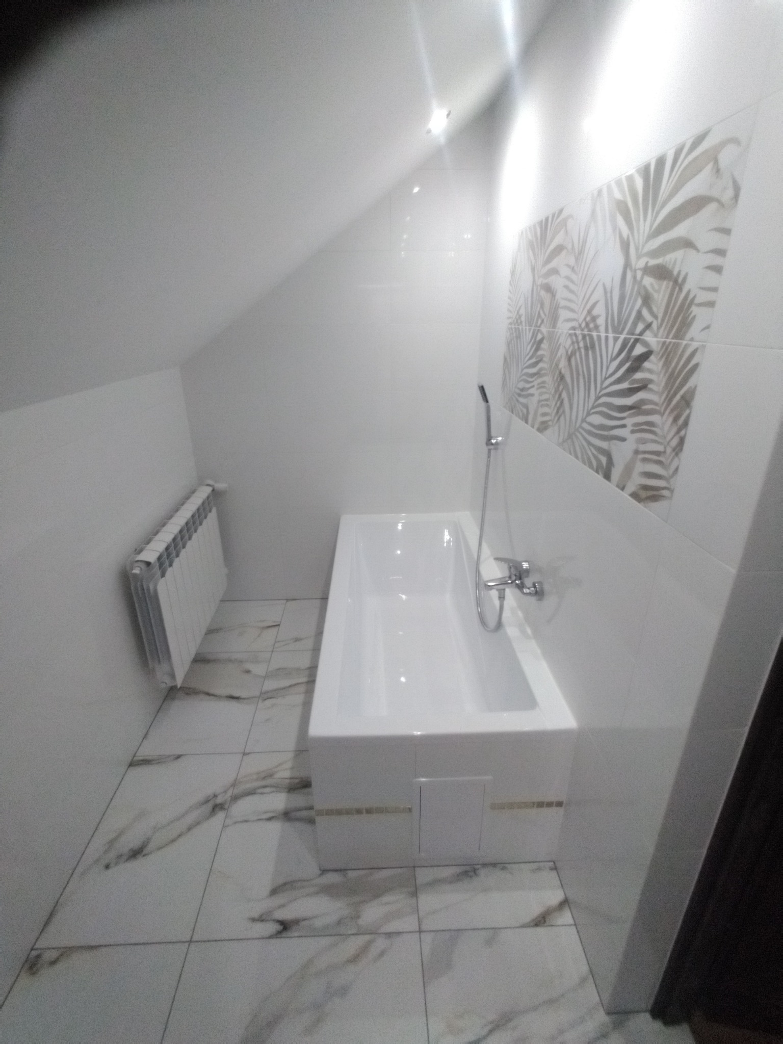 Biała wanna z baterią i prysznicem w łazience z białymi płytkami i dekoracyjnym pasem z motywem liści, grzejnik na ścianie, podłoga z marmurowymi płytkami.