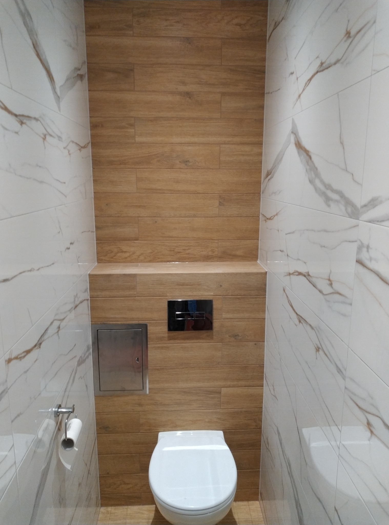 Wąska toaleta z białymi marmurkowymi płytkami na ścianach bocznych i drewnopodobnymi płytkami na ścianie tylnej, z podtynkową spłuczką, klapą rewizyjną oraz uchwytem na papier toaletowy.