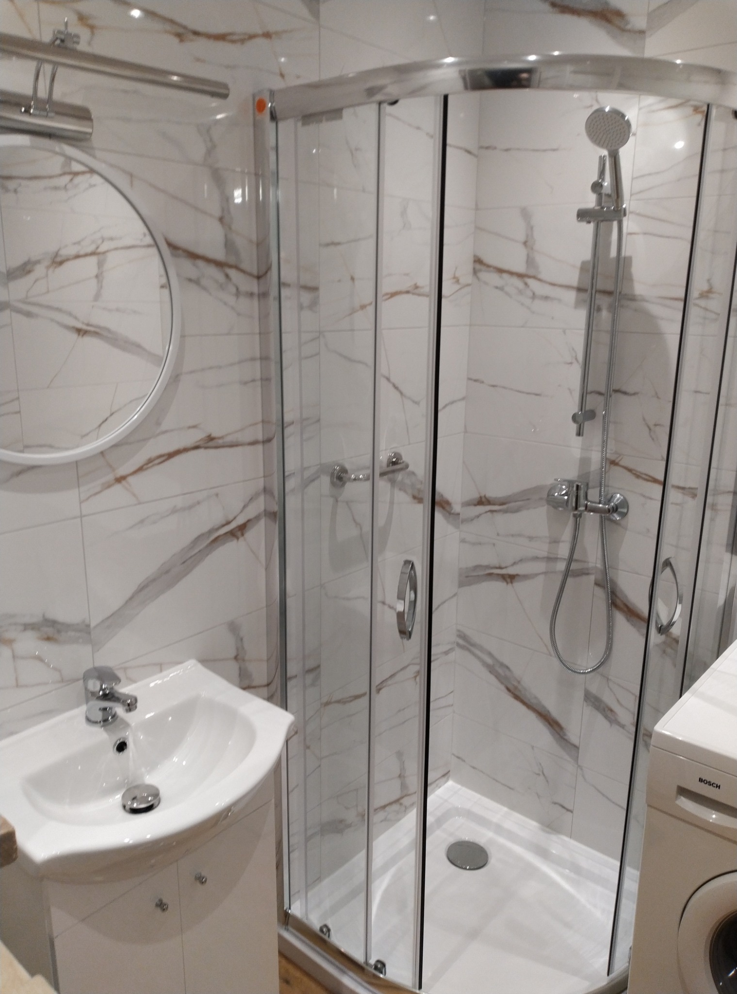 Nowoczesna łazienka z kabiną prysznicową, umywalką na szafce i okrągłym lustrem, wykończona białymi płytkami z brązowym marmurowym wzorem.