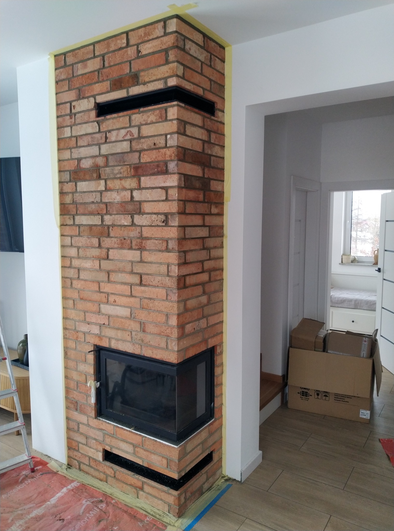 Murowany kominek z cegły w trakcie budowy, zabezpieczony taśmą malarską, z widocznym wkładem kominkowym i otworami wentylacyjnymi, na podłodze folia ochronna.