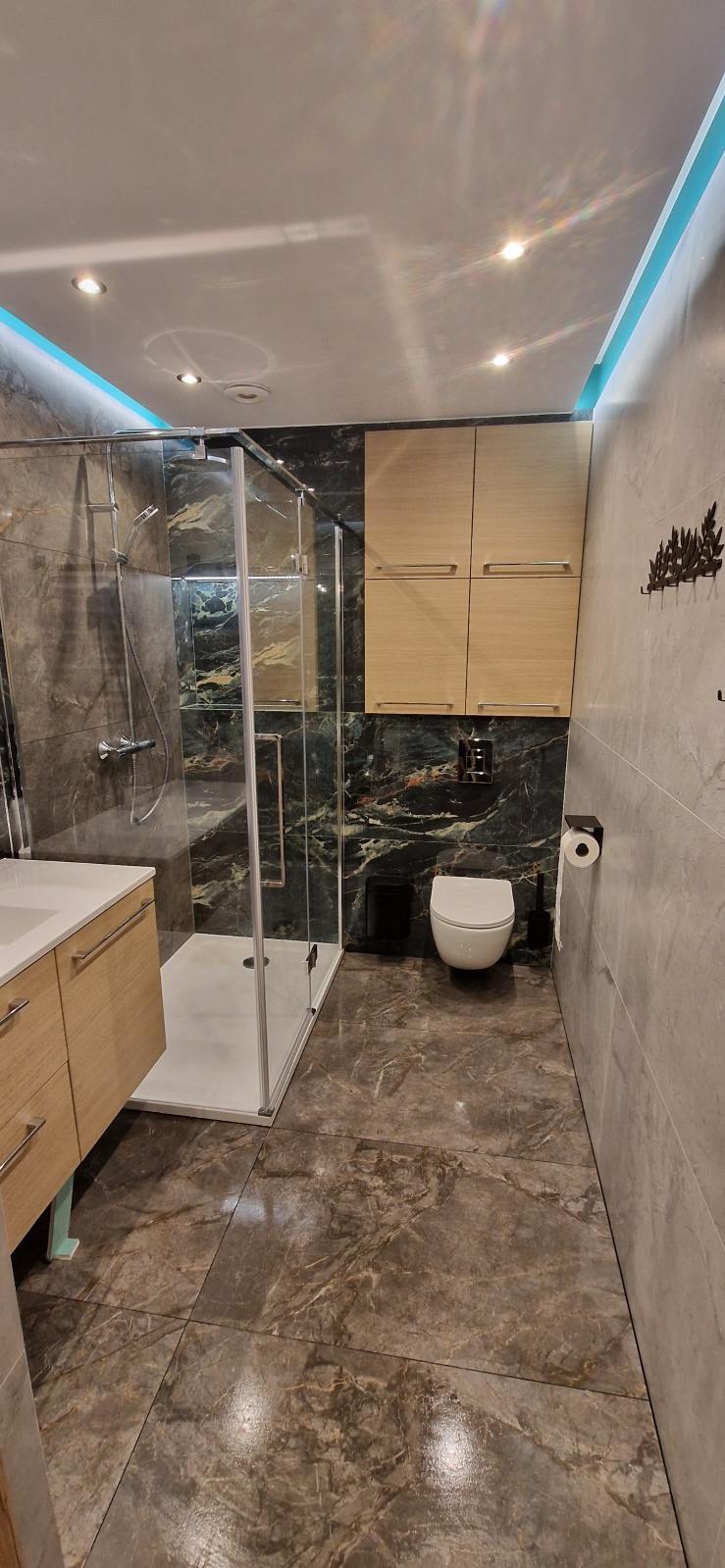 Nowoczesna łazienka z kabiną prysznicową typu walk-in, marmurowymi płytkami na ścianach i podłodze, oraz drewnianymi szafkami wiszącymi nad toaletą.