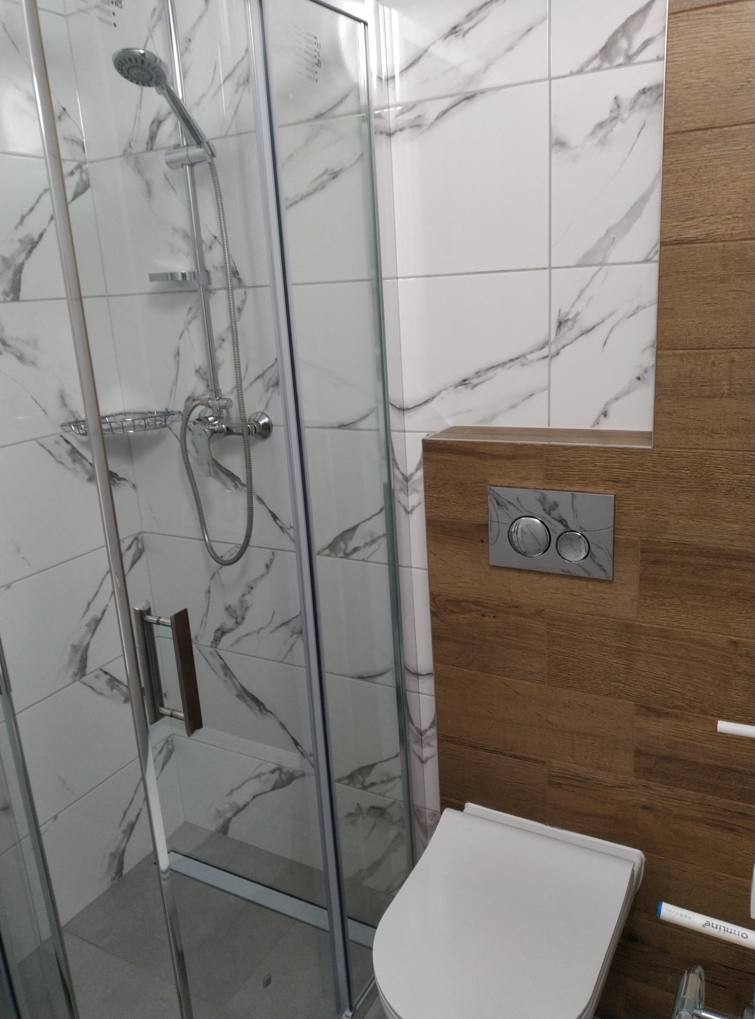 Nowoczesna łazienka po remoncie: kabina prysznicowa z marmurowymi płytkami, toaleta z drewnianą obudową i przyciskiem spłukiwania z efektem marmuru.