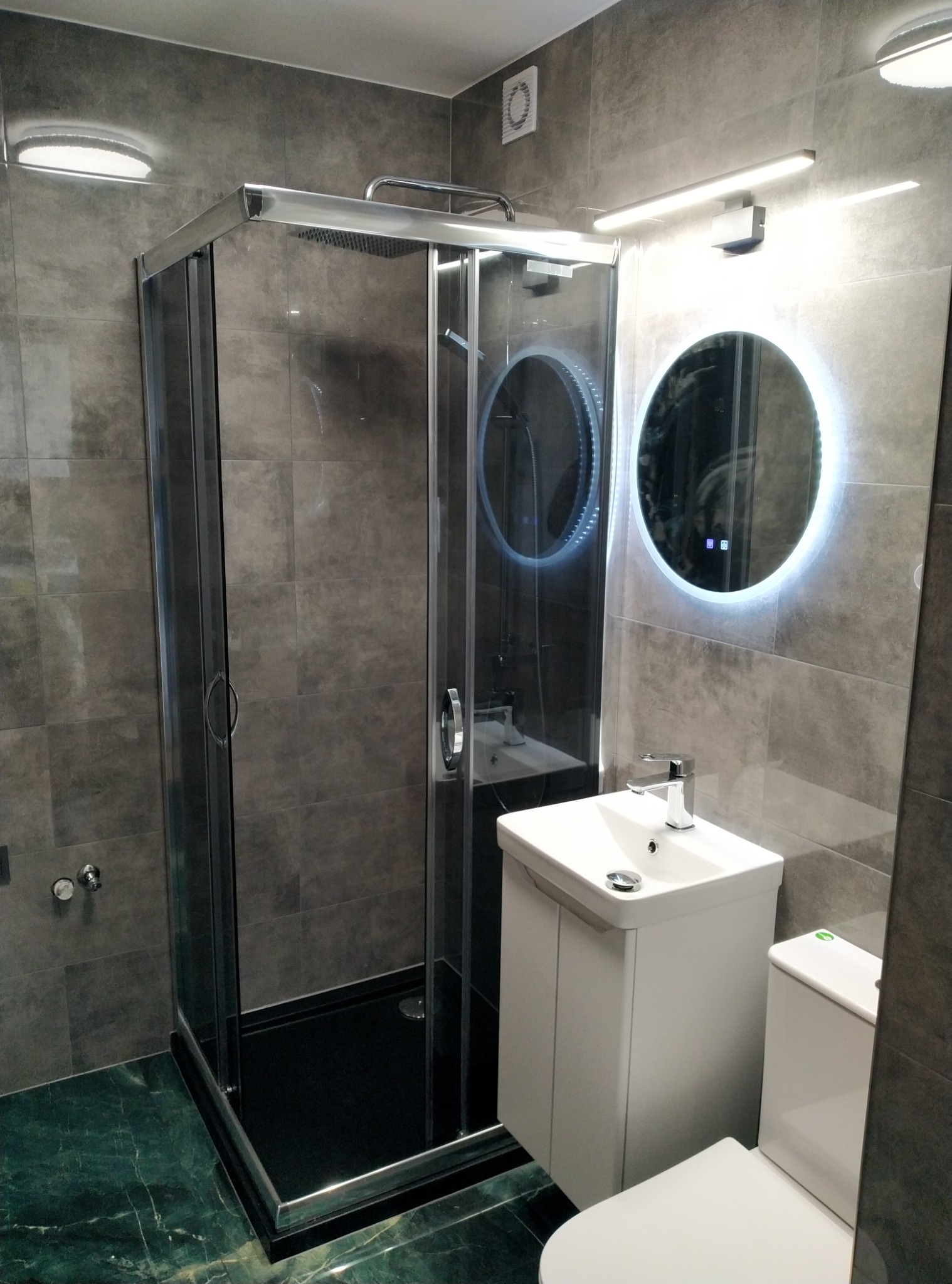 Nowoczesna łazienka z kabiną prysznicową, umywalką z szafką i toaletą. Szare płytki i okrągłe lustro z podświetleniem LED. Marmurowa podłoga.