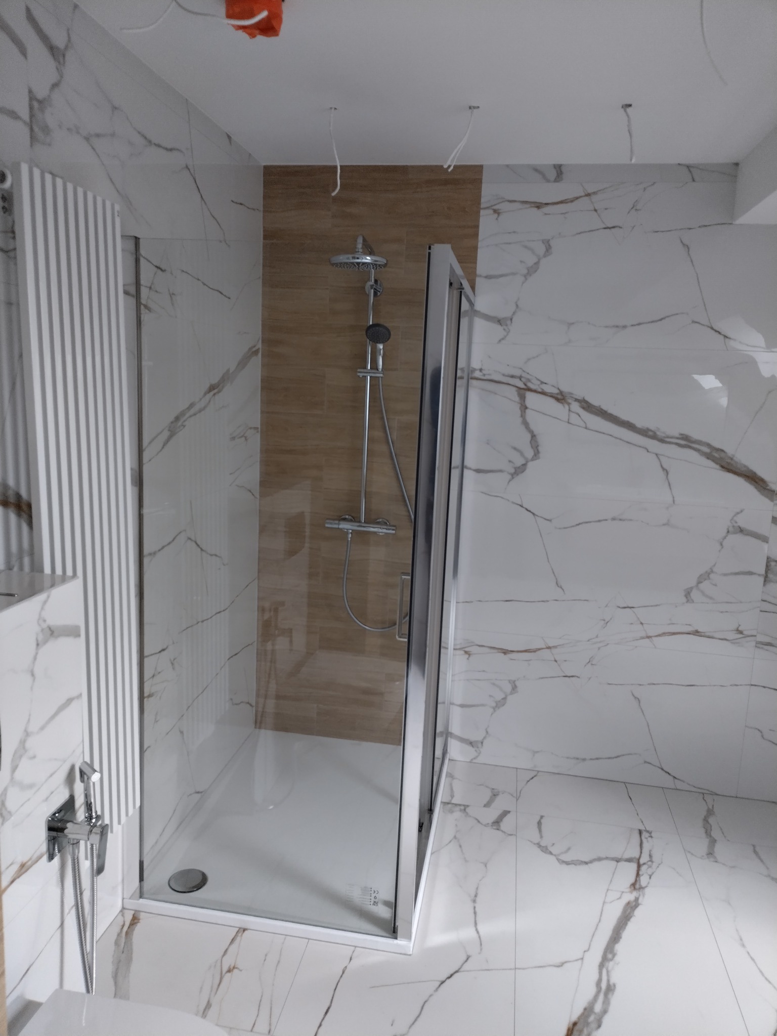 Nowoczesna kabina prysznicowa z deszczownicą, wykończona płytkami imitującymi marmur. Widoczne elementy instalacji elektrycznej na suficie.