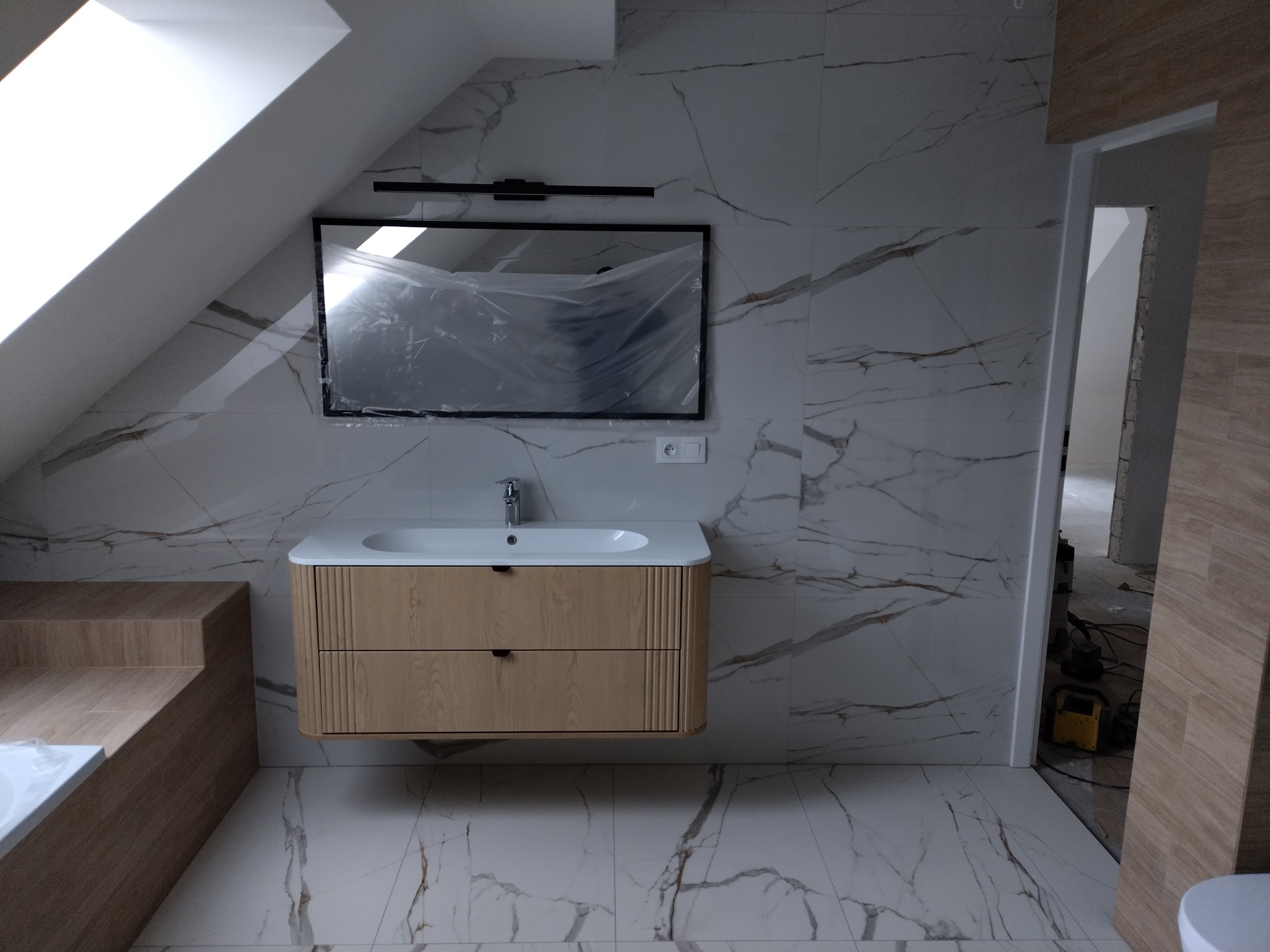 Wykończona łazienka na poddaszu z marmurowymi płytkami, drewnianą szafką pod umywalką i lustrem w folii ochronnej. Widoczny fragment wanny i wejście do pomieszczenia.