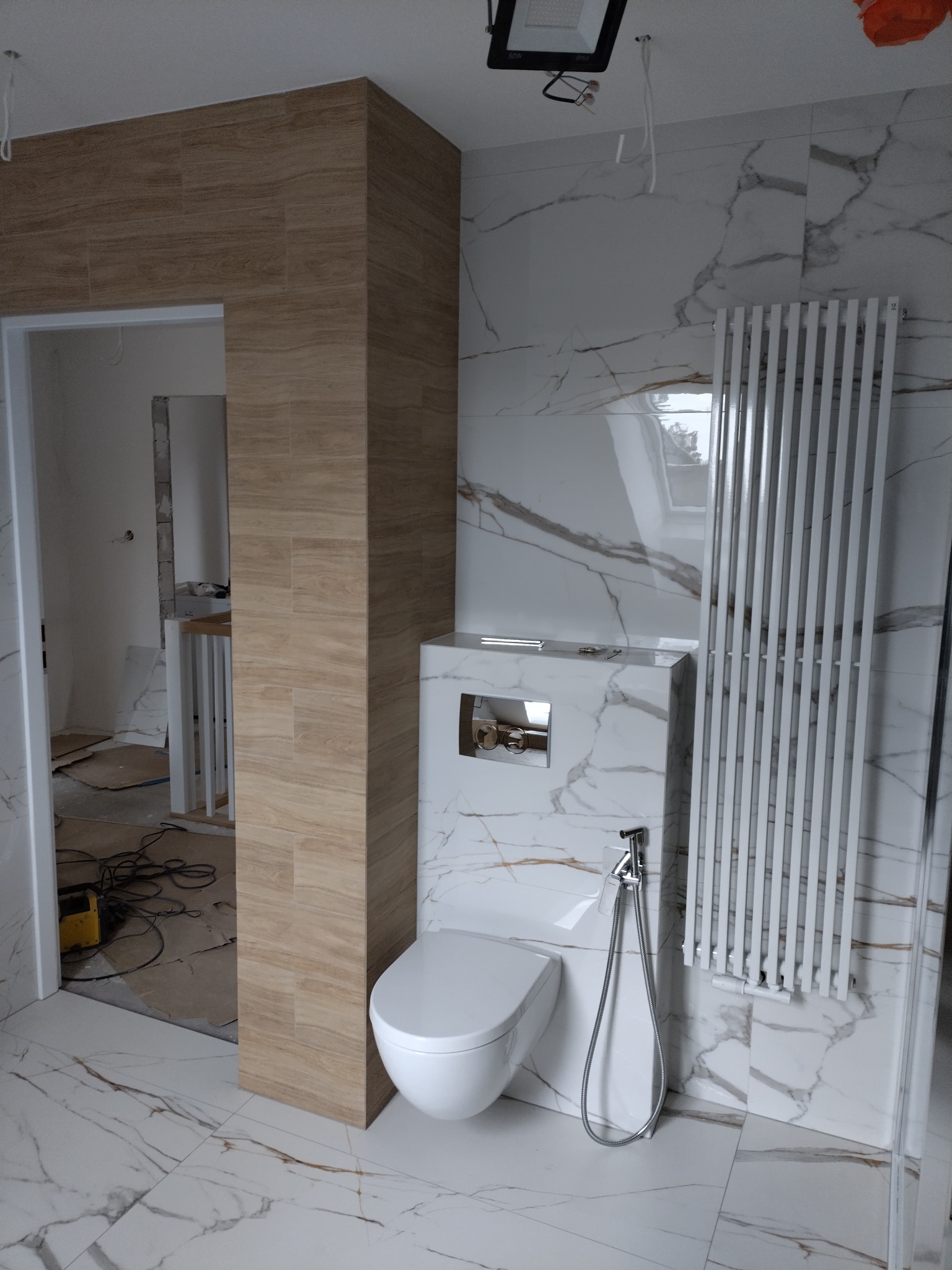 Wykończona łazienka z toaletą podwieszaną, grzejnikiem drabinkowym i płytkami imitującymi marmur. Widoczne niedokończone pomieszczenie obok.
