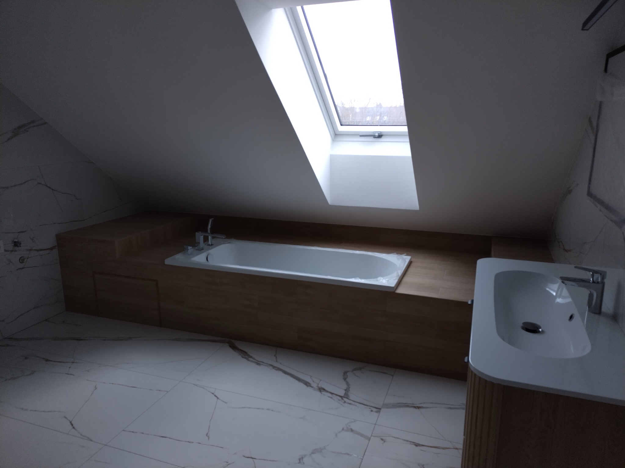 Nowoczesna łazienka na poddaszu z oknem dachowym, białą wanną w zabudowie z drewnopodobnych paneli i marmurową posadzką. Umywalka nablatowa z szafką.