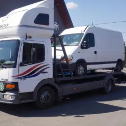 Laweta Mercedes-Benz Atego z białym busem na platformie, widok z boku na tle budynku i zieleni. Na pojeździe napis 'Pomoc Drogowa'.