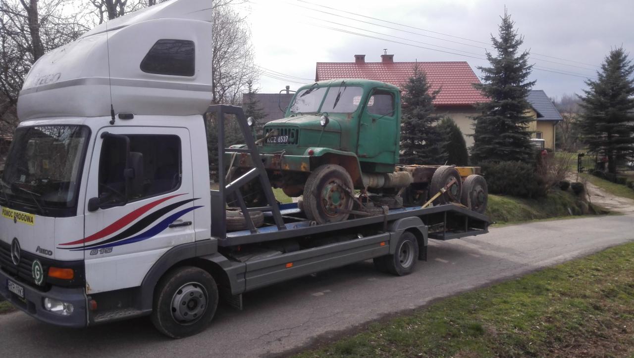 Laweta z białym kabiną transportuje stary, zielony samochód ciężarowy zardzewiały na platformie. W tle domy jednorodzinne i drzewa.