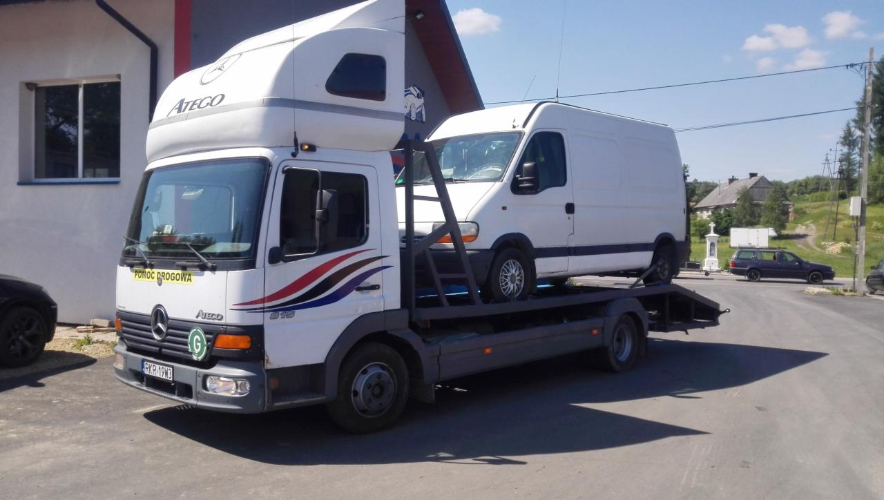 Laweta Mercedes-Benz Atego z białym busem na platformie, widok z boku na tle budynku i zieleni. Na pojeździe napis 'Pomoc Drogowa'.