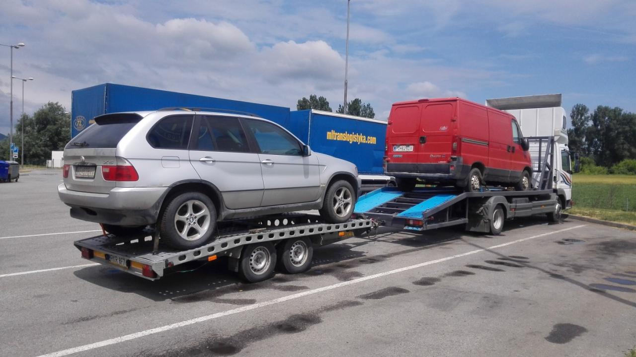 Laweta z srebrnym BMW X5 i czerwonym busem na niebieskiej platformie, zaparkowana na parkingu z plamami, w tle błękitne niebo.