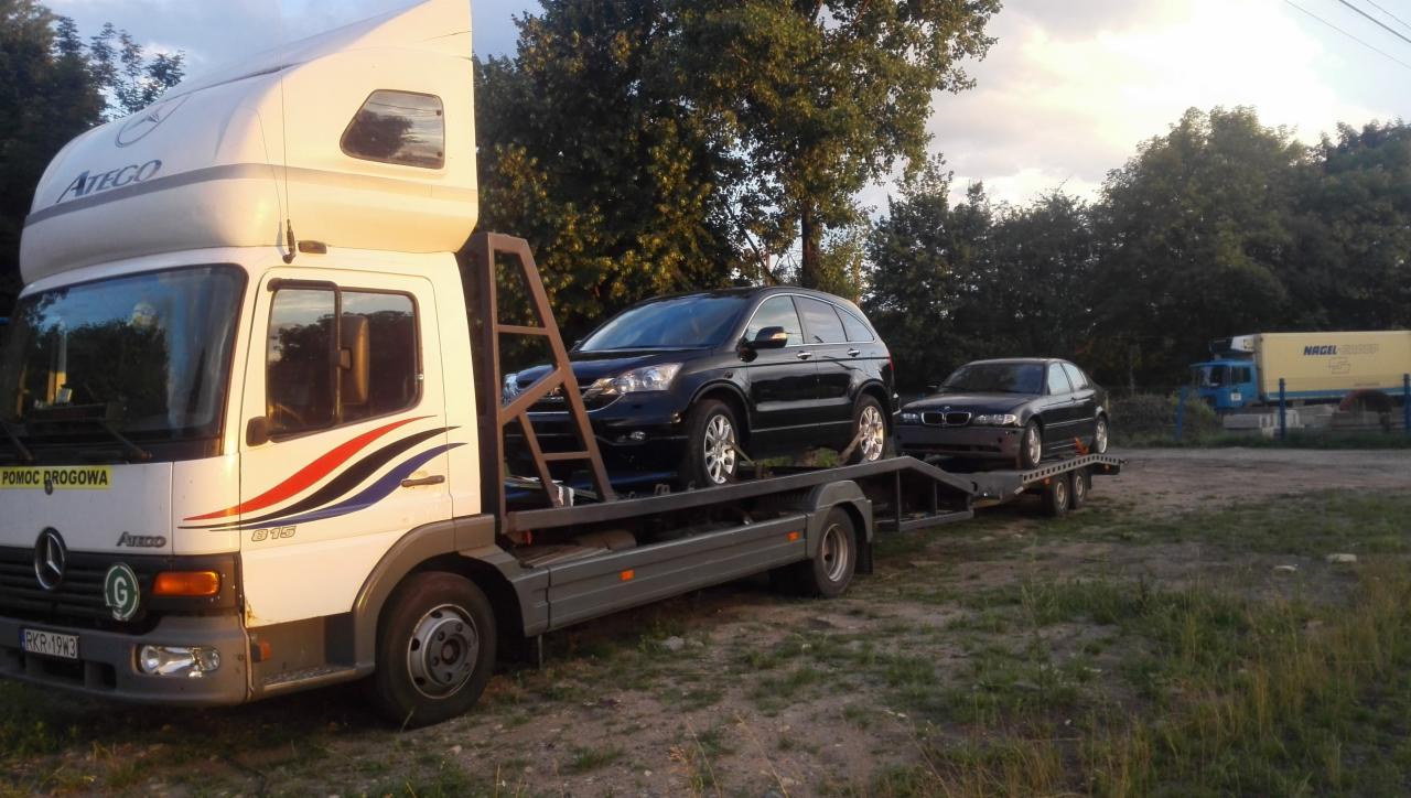 Laweta Mercedes Atego z dwoma czarnymi samochodami na platformie, zaparkowana na trawiastym terenie, w tle drzewa i budynki.
