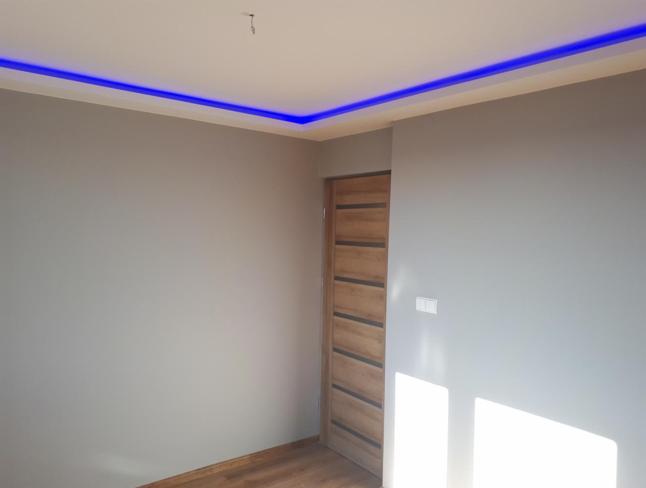 Szare wnętrze z drewnianą podłogą i drzwiami, oświetlone niebieskim paskiem LED pod sufitem, widoczne gniazdko elektryczne na ścianie.