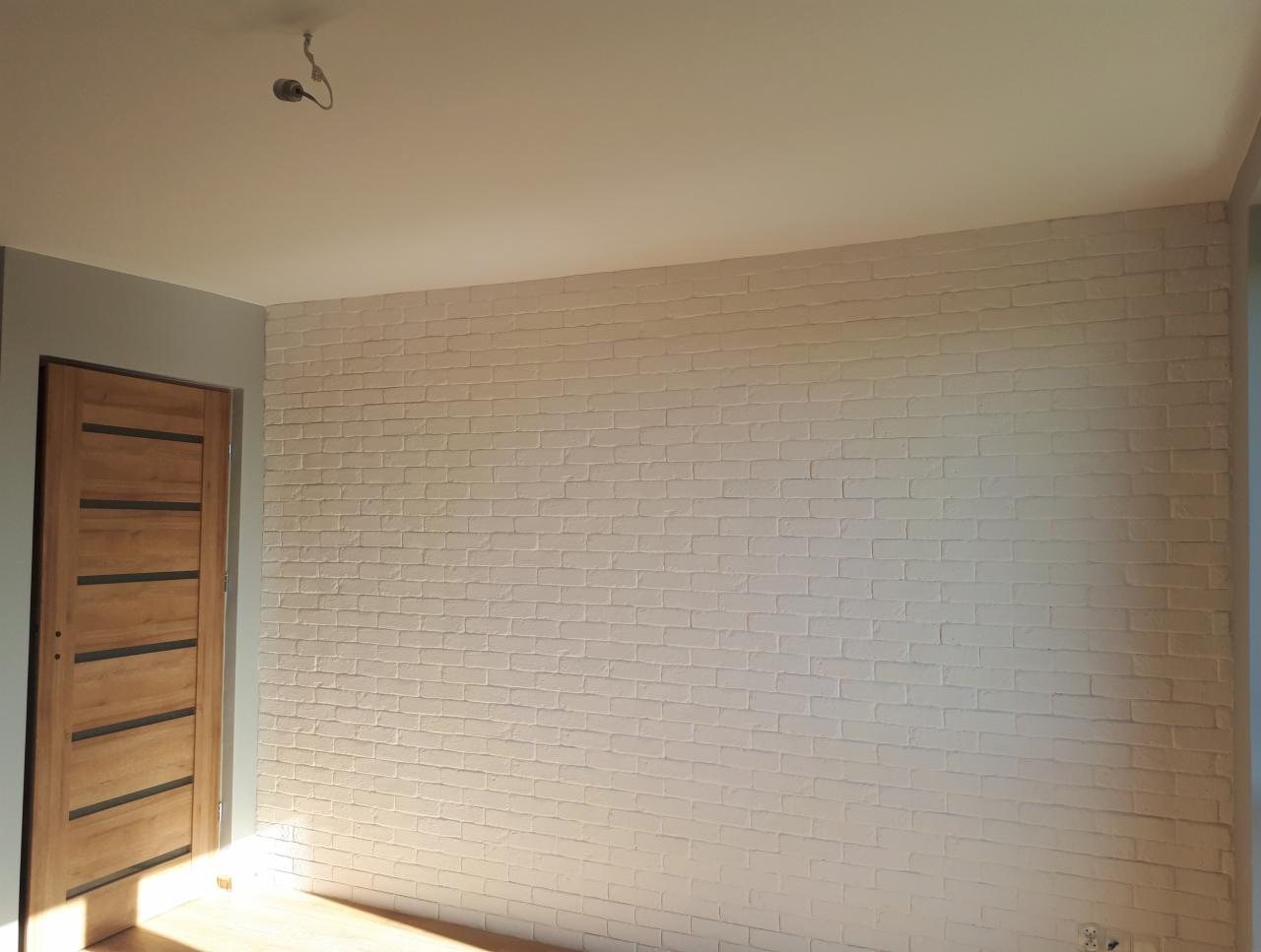 Ściana z cegły pomalowana na biało z widoczną strukturą, drewniane drzwi z poziomymi czarnymi paskami, jasne drewniane podłogi, widoczne gniazdko elektryczne.