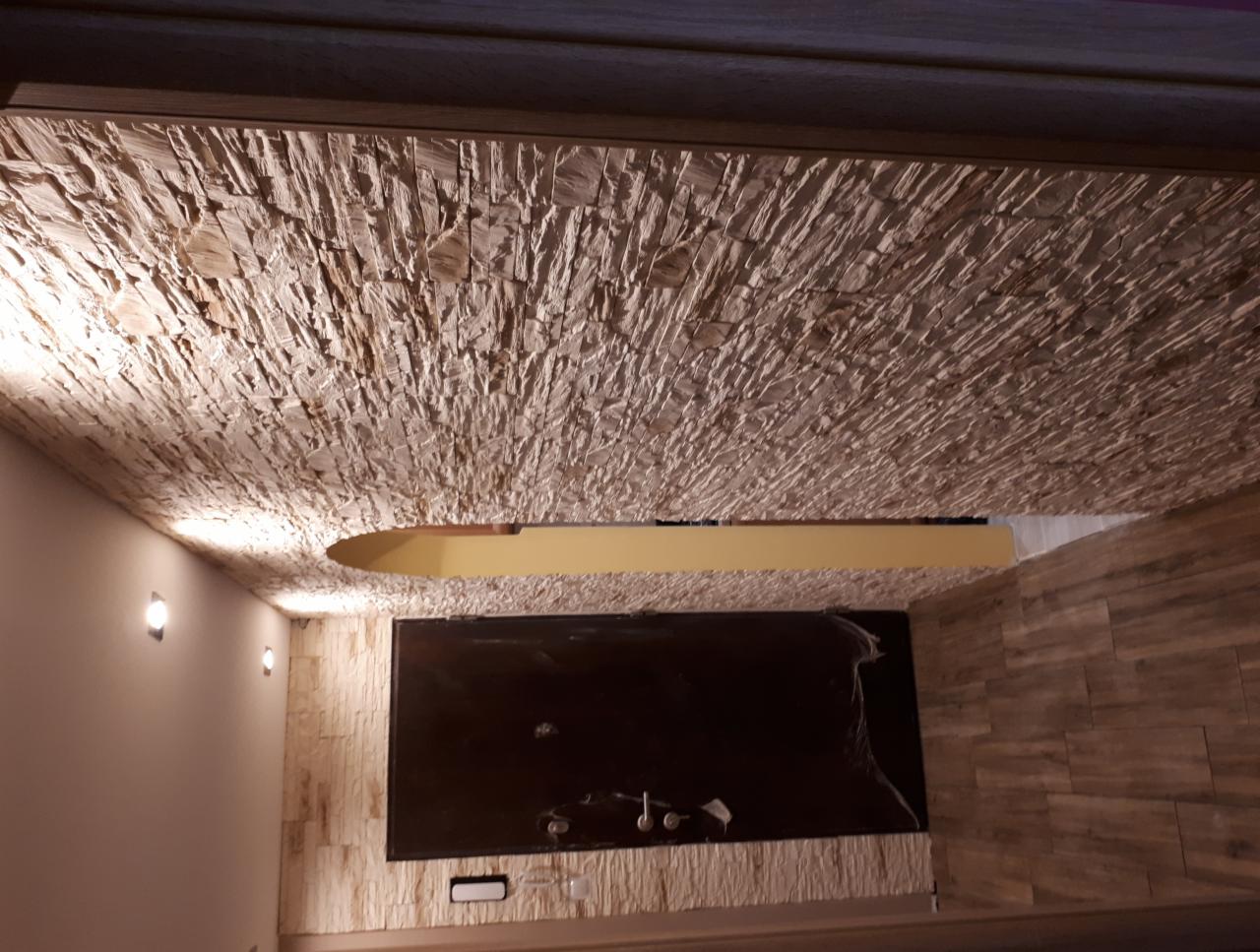 Kamienna okładzina sufitowa z oświetleniem punktowym, uzupełniona ścianą z płytek imitujących drewno i czarnym panelem na ścianie.