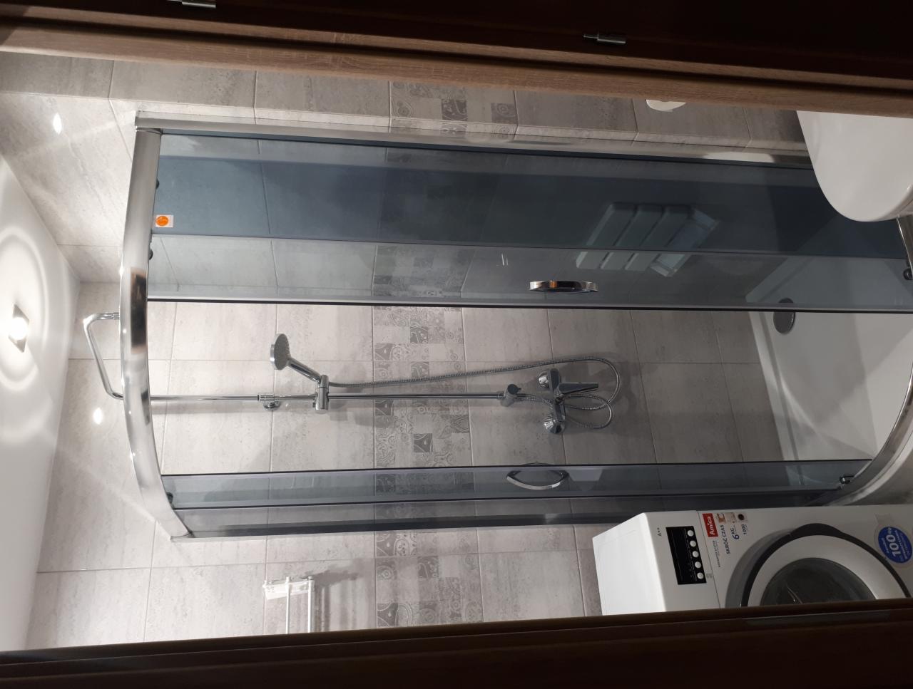 Szklana kabina prysznicowa z przesuwnymi drzwiami w łazience z szaro-beżowymi płytkami, widoczny fragment umywalki, pralka i drewniana rama drzwi.