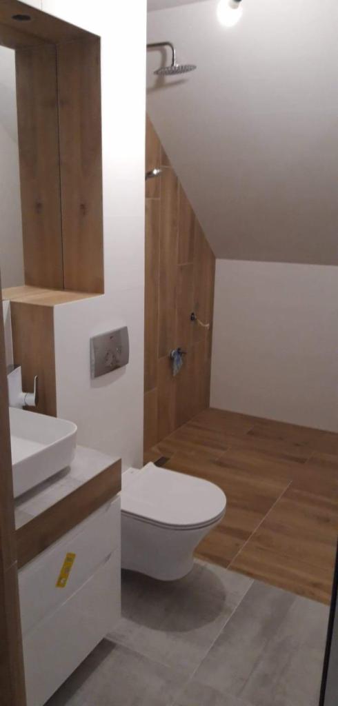 Nowoczesna łazienka na poddaszu z drewnopodobnymi płytkami na ścianie prysznica i podłodze, białą ceramiką sanitarną i szarą podłogą.