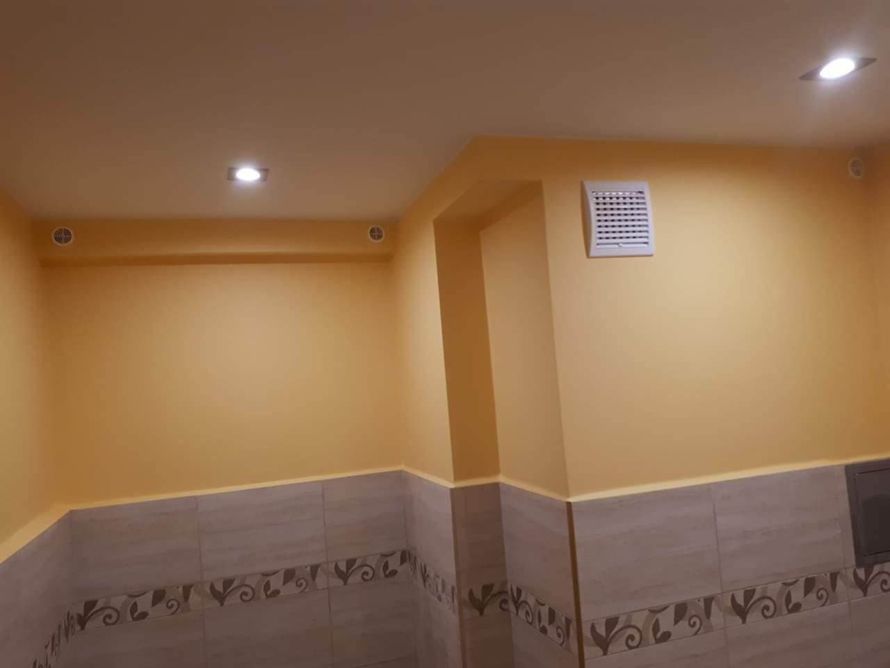 Żółta ściana w łazience z kremowymi płytkami dekoracyjnymi u dołu, kratka wentylacyjna na ścianie i oświetlenie sufitowe.