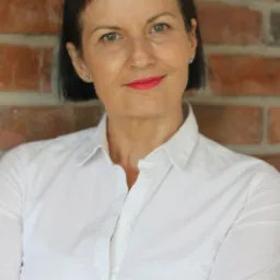 Portret kobiety z krótkimi, ciemnymi włosami i różowymi ustami, ubrana w białą koszulę, stojąca na tle ceglanej ściany. Uśmiecha się z założonymi rękami.