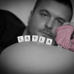 N.O-wska Fotografia - Czarno-białe zdjęcie ciężarnej kobiety z imieniem 'Layla' ułożonym z płytek Scrabble na brzuchu, obok różowych skarpetek niemowlęcych trzymanych przez mężczyznę w tle.