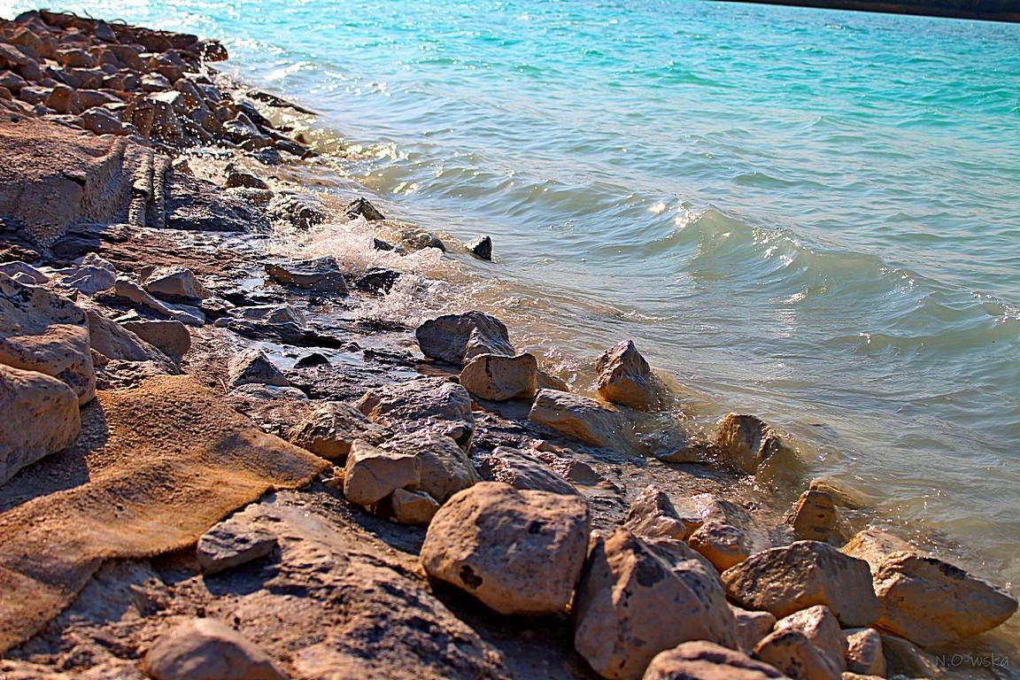Brzeg z kamieniami i turkusową wodą, delikatne fale rozbijają się o skały.