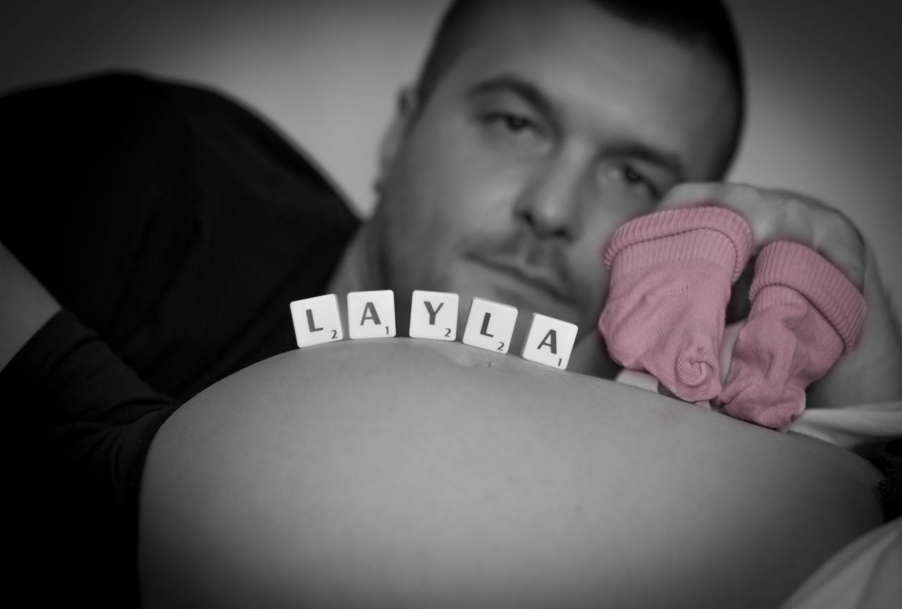 Czarno-białe zdjęcie ciężarnej kobiety z imieniem 'Layla' ułożonym z płytek Scrabble na brzuchu, obok różowych skarpetek niemowlęcych trzymanych przez mężczyznę w tle.