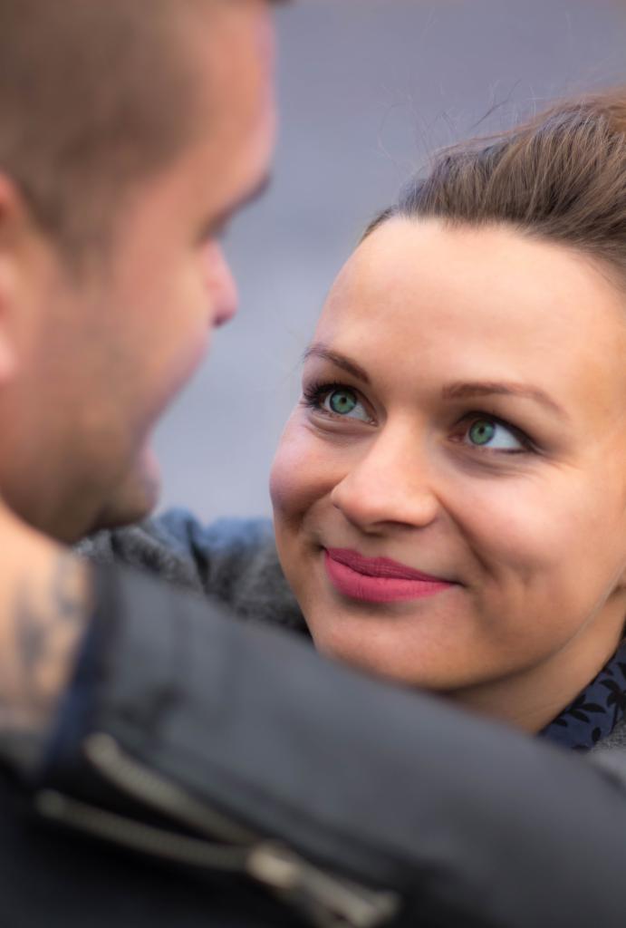 Uśmiechnięta kobieta z zielonymi oczami spogląda w górę na mężczyznę, widoczny fragment jego twarzy i tatuaż na ramieniu, miękkie oświetlenie.