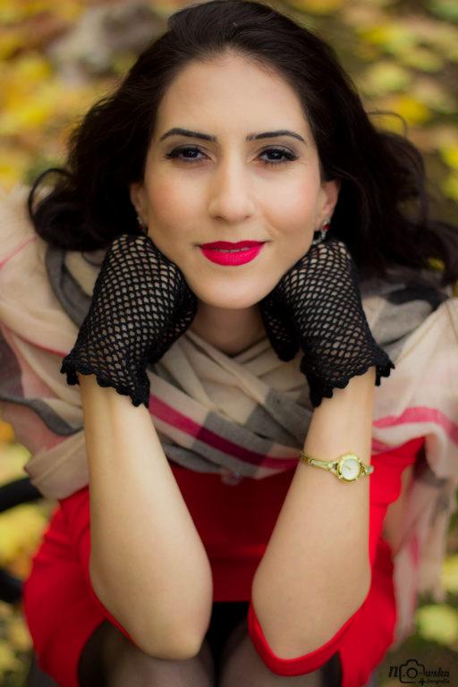 Portret kobiety w czerwonej sukience, czarnych rękawiczkach z siatki i wzorzystym szaliku na tle opadłych jesiennych liści, z delikatnym uśmiechem i wyrazistym makijażem.