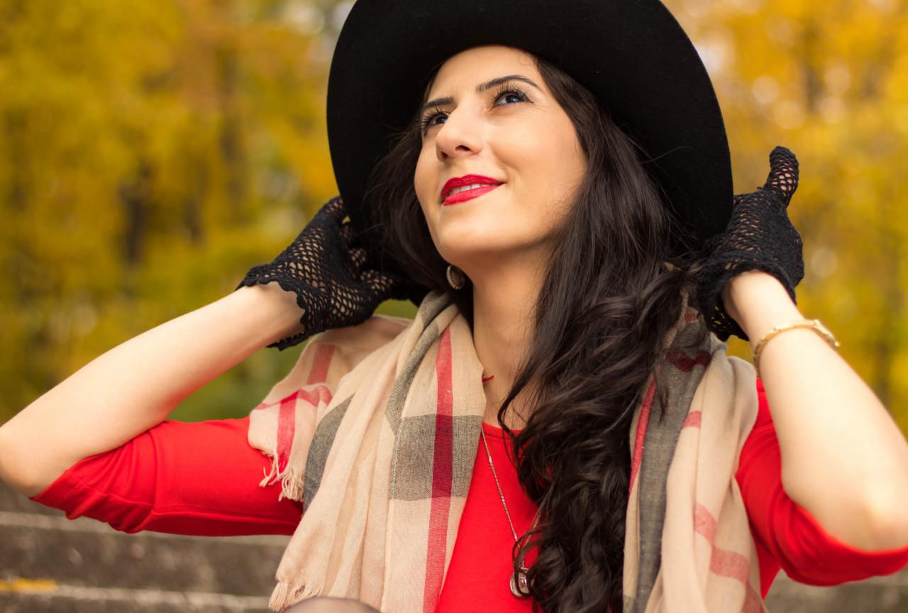 Portret kobiety w czarnym kapeluszu, czerwonej sukience i ażurowych rękawiczkach, spoglądającej w górę na tle jesiennych drzew.