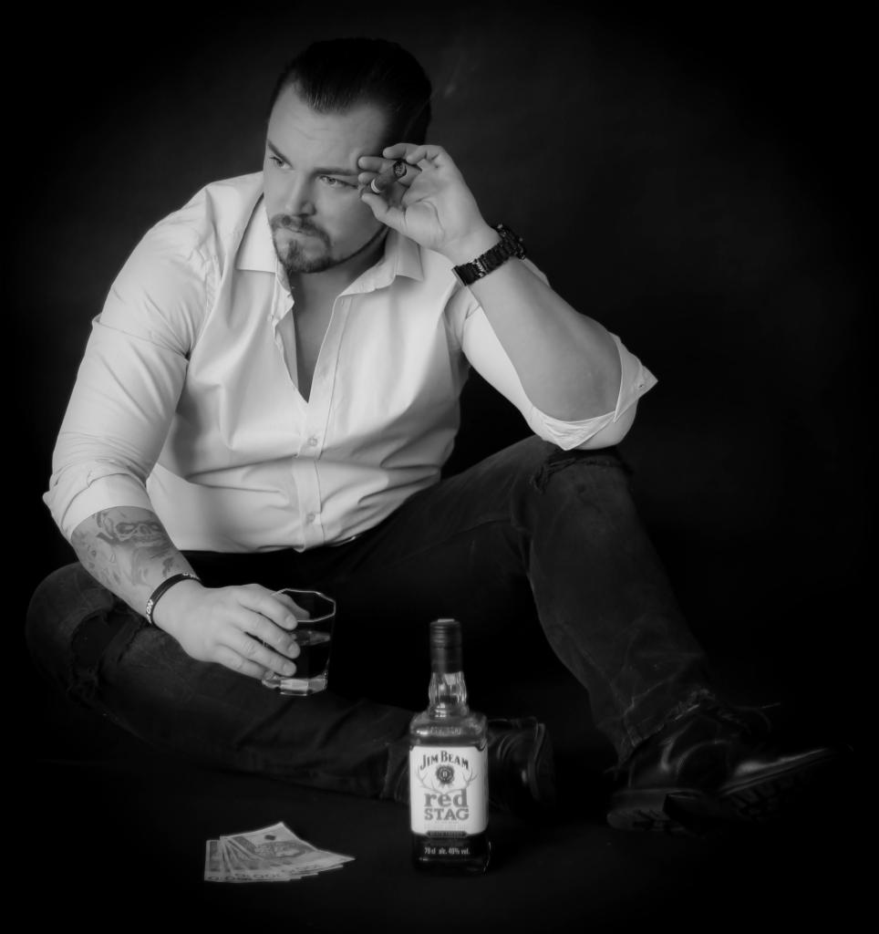 Mężczyzna w rozpiętej białej koszuli, siedzący na podłodze, trzyma szklankę z napojem, obok butelka Jim Beam Red Stag i banknoty, czarno-biała fotografia.