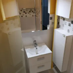 Biała umywalka z szafką z dwoma szufladami i chromowaną baterią w wyremontowanej łazience z białymi płytkami z dekoracyjnym motywem i żółtymi akcentami na ścianach, widok na lustro z oświetleniem.