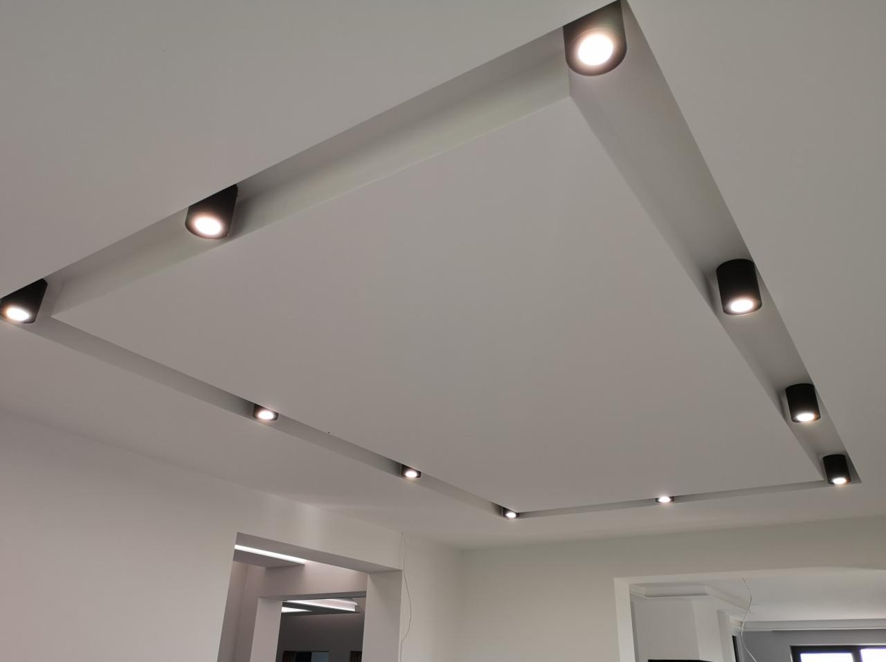 Nowoczesny sufit podwieszany z wbudowanymi czarnymi reflektorami LED, tworzący geometryczny wzór oświetleniowy w jasnym wnętrzu.