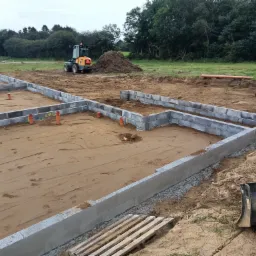 Wykop pod fundamenty z ułożonymi bloczkami betonowymi, widoczne rury kanalizacyjne i dwa mini-koparki na placu budowy.