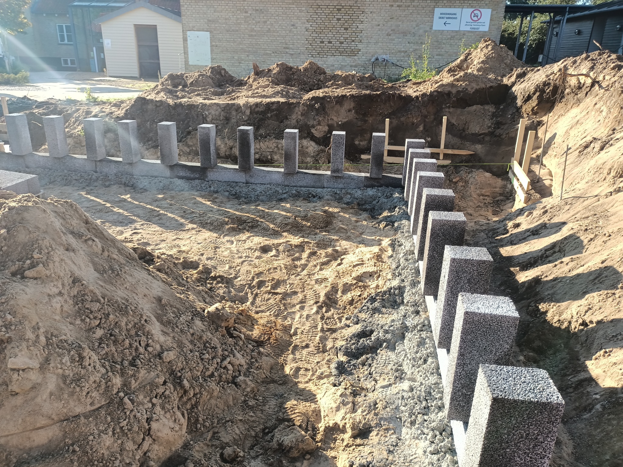 Budowa fundamentów: rów z ułożonymi blokami betonowymi, widoczne ślady opon w piasku i drewniane szalunki w tle.