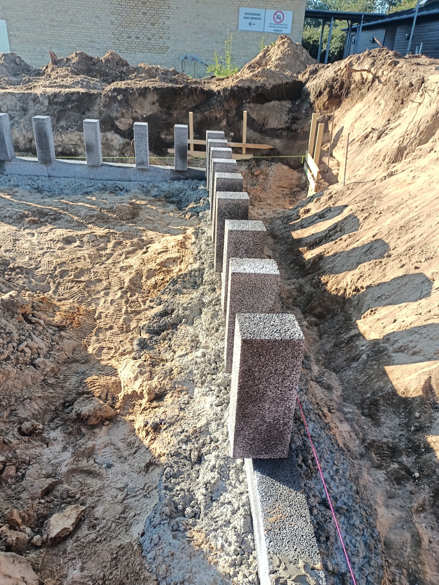 Wykop pod fundamenty z częściowo wzniesioną ścianą z bloczków betonowych, widoczna różowa nić wyznaczająca linię muru.