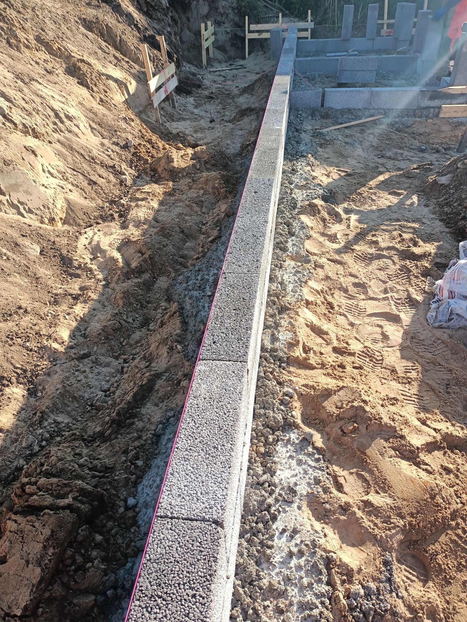 Wykop pod fundament z ułożonym pierwszym rzędem bloczków betonowych, widoczne ślady opon i stopy w piasku, teren budowy.