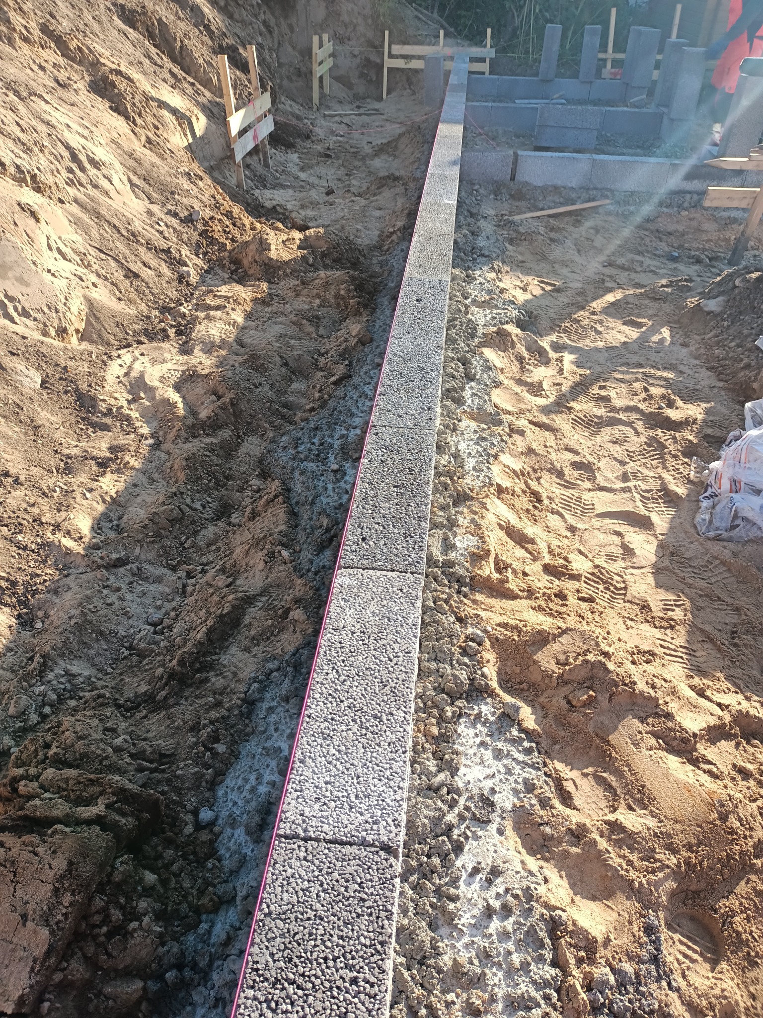 Wykop pod fundamenty z ułożonym pierwszym rzędem bloczków betonowych, widoczne ślady opon i stopy na piasku.