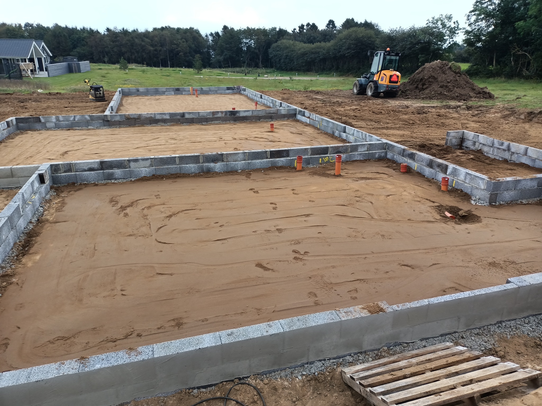 Wykop pod fundamenty domu z częściowo wymurowanymi ścianami z szarych bloczków betonowych, widoczne pomarańczowe rury kanalizacyjne i sprzęt budowlany w tle.