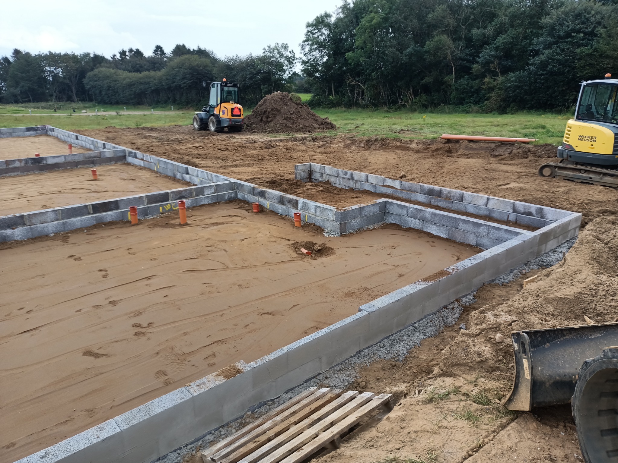 Wykop pod fundamenty z ułożonymi bloczkami betonowymi, widoczne rury kanalizacyjne i dwa mini-koparki na placu budowy.