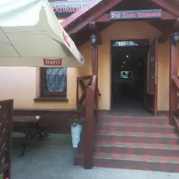 Wejście do pubu Stara Winiarnia z zewnętrznym stolikiem pod parasolem, widoczne schody z płytek i kwiaty w donicach.